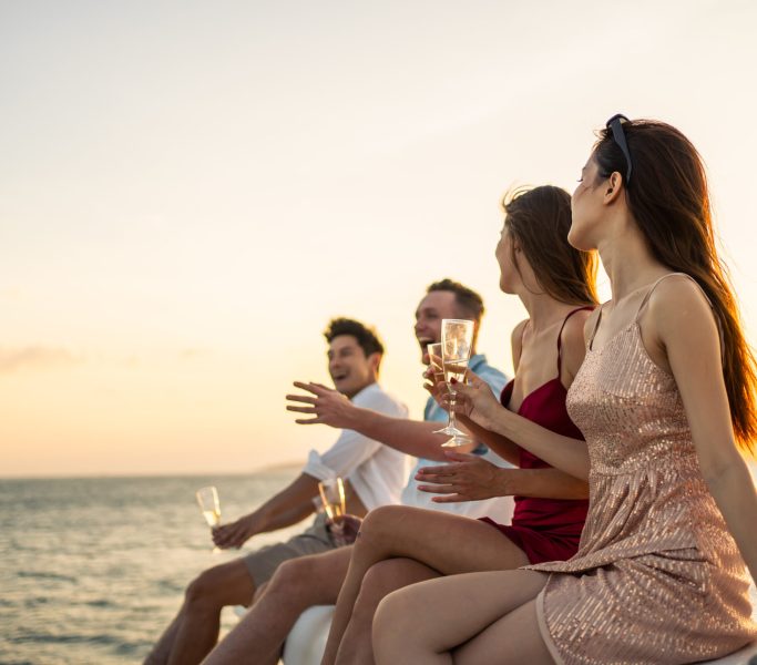 Group of diverse friends drink champagne while having a party in yacht. Attractive young men and women hanging out, celebrating holiday vacation trip while catamaran boat sailing during summer sunset.