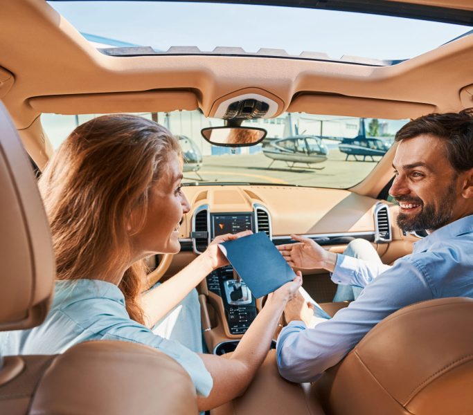 Merry lady presenting gift certificate for helicopter tour to smiling pleased young man in automobile